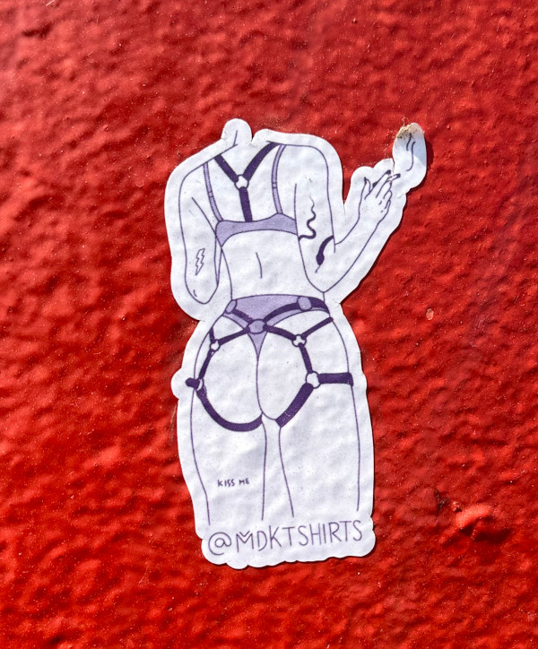 Sticker by @MDKTSHIRTS on a red background, showing the back of a woman with tattoos and harness while smoking